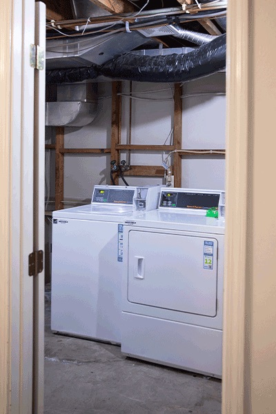 A view into a spacious laundry room with a clean, white washer and dryer. There are exposed beams and pipes.